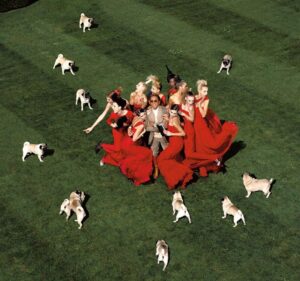 Valentino the designer in a green field surrounded by women in gorgeous red g owns and a field of pugs surrounding them 